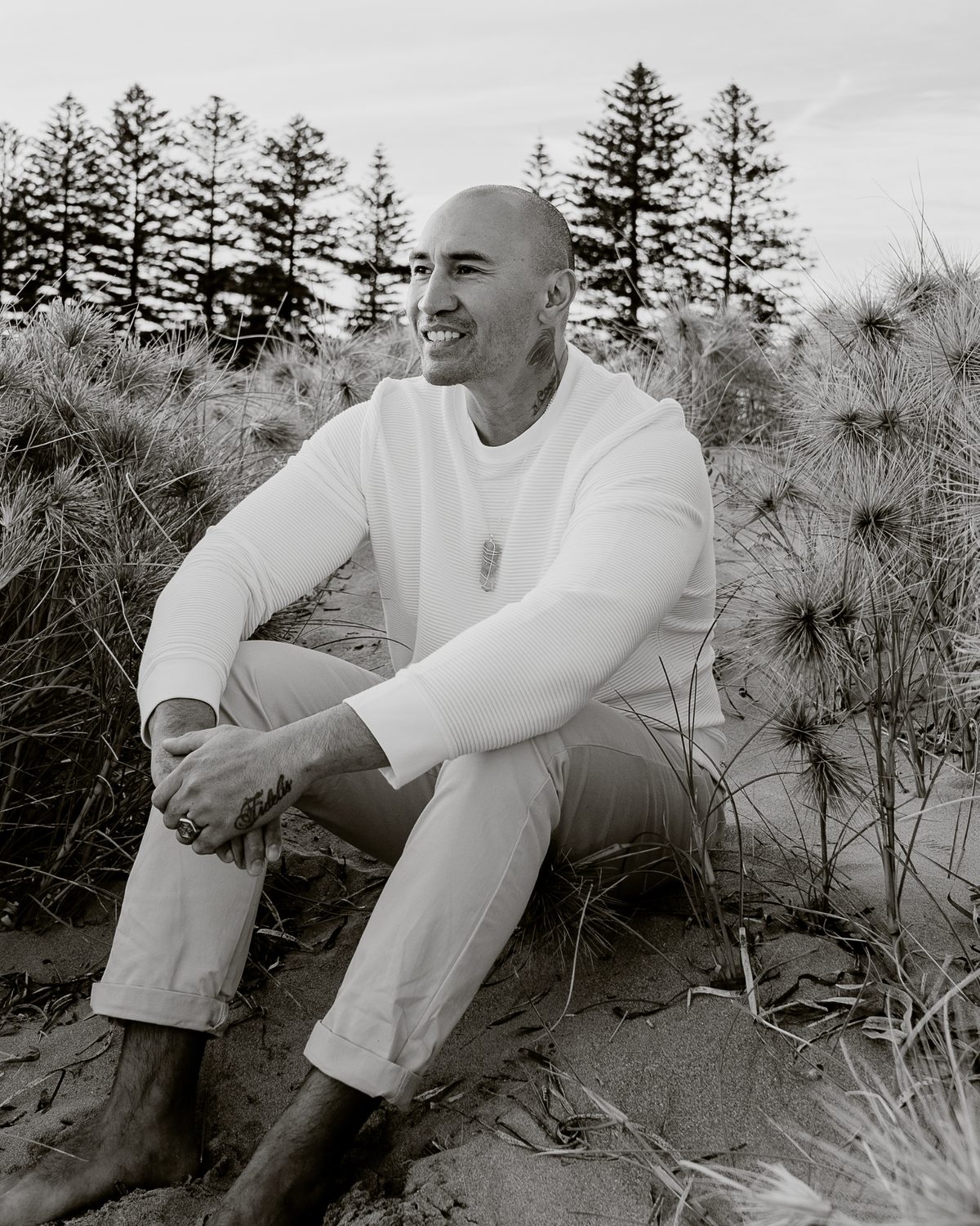 LJ Silulu seated among dune grass with pine trees behind him, smiling and looking off to one side, in a black and white photograph.