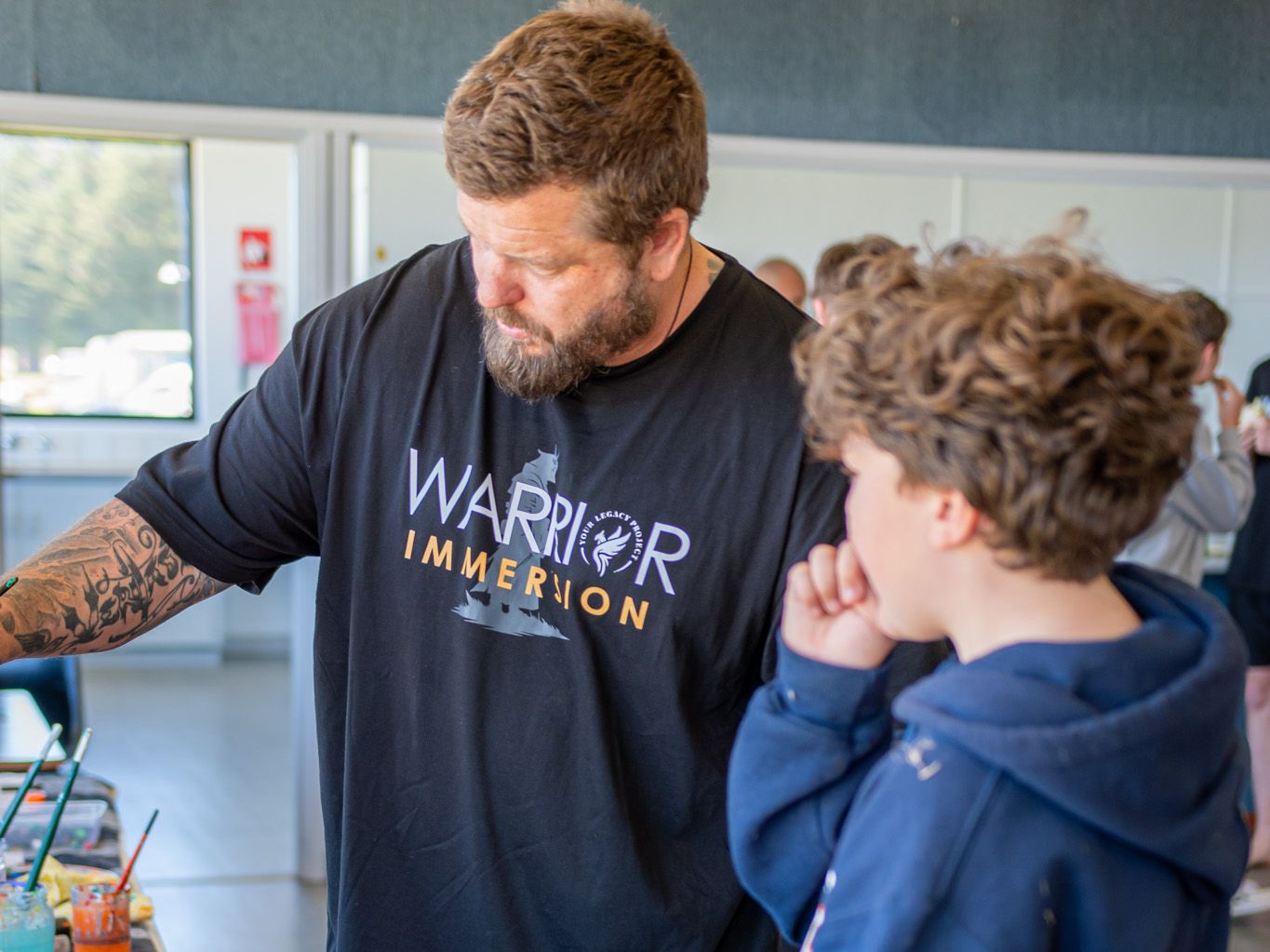 A mentor leaning over a painting table, looking down at the work, with a teenage boy in a navy hoodie watching beside him in a bright hall.
