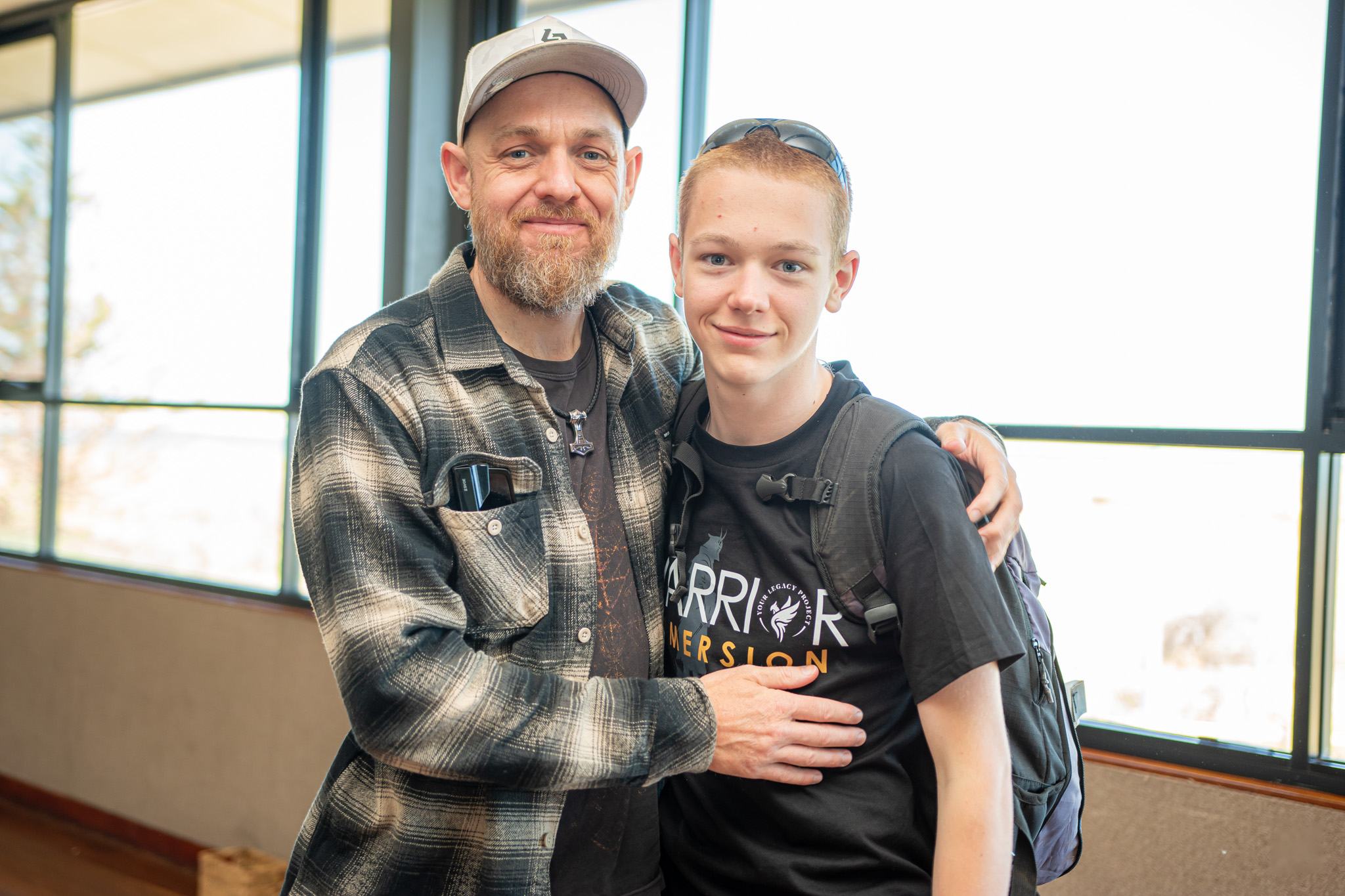 A mentor and a teenage boy standing side by side, smiling at the camera, the mentor with an arm around the boy's shoulder.