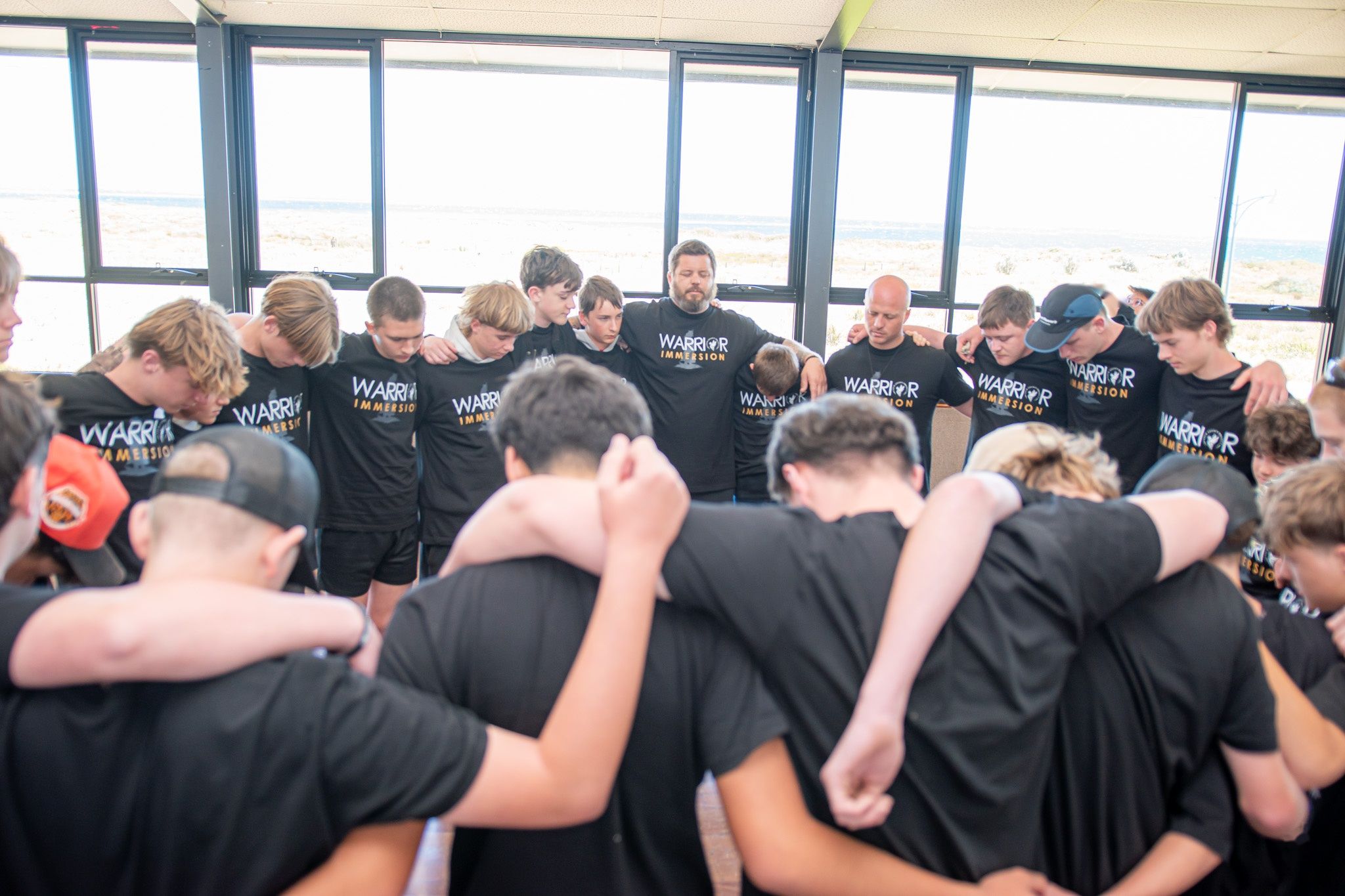 A group of teenage boys and their mentors standing in a circle with arms around each other and heads bowed, in a bright room overlooking the coast.