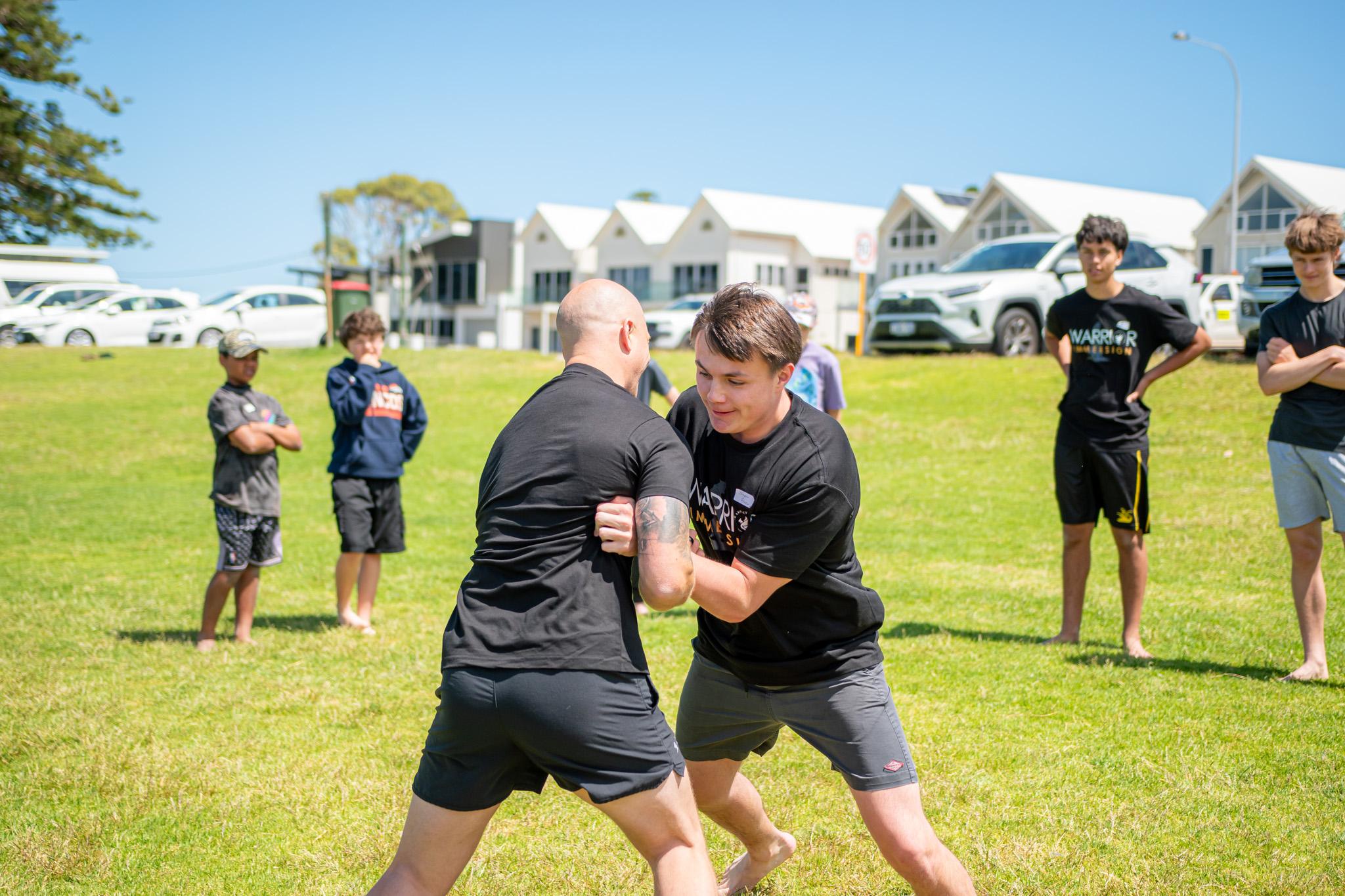 A mentor and a teenage boy grappling in a friendly practice contest on a grass field while other boys watch on.