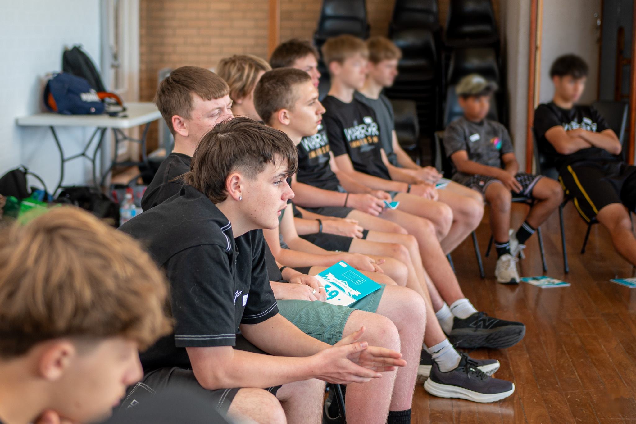 A group of teenage boys sitting in a row on chairs in a hall, listening during a workshop.
